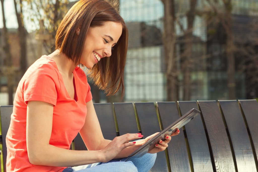 Young woman sits in a park on a wooden bench and using tablet